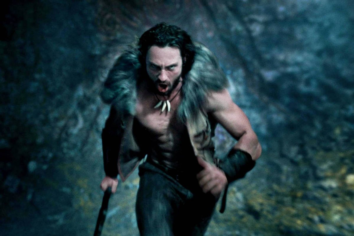 Um homem musculoso e sem camisa corre em um ambiente escuro e rochoso. Ele usa uma pele de animal nos ombros e tem um colar de dentes ou ossos. Sua expressão é intensa e determinada, e ele segura um bastão com uma das mãos.