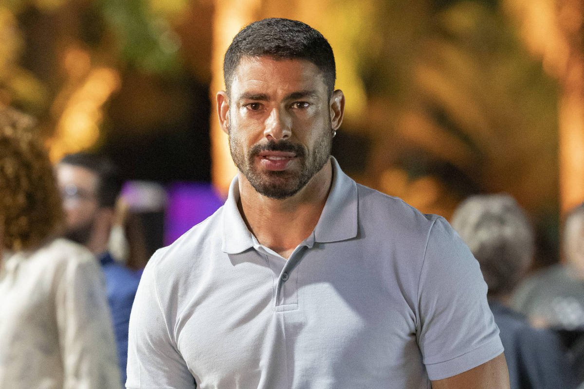 Um homem está em um evento ao ar livre, usando uma camisa polo cinza e shorts pretos. Ele tem cabelo curto e barba bem aparada, e está posando com uma expressão séria. Ao fundo, há pessoas conversando e árvores, sugerindo um ambiente social.