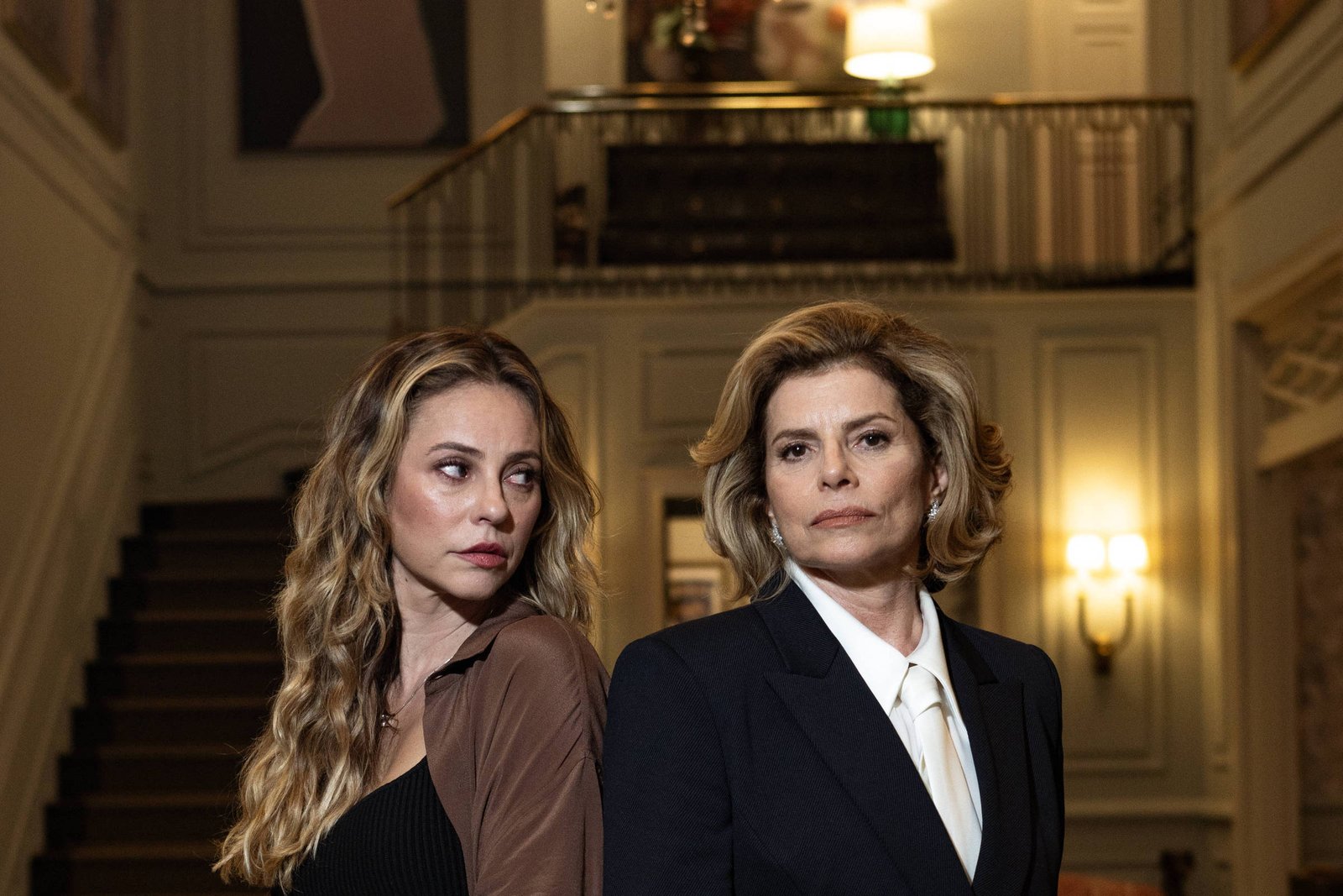 A imagem mostra duas mulheres em um ambiente interno sofisticado, com escadas ao fundo e um lustre pendurado. A mulher à esquerda tem cabelo longo e ondulado, vestindo uma blusa preta e uma jaqueta marrom. A mulher à direita tem cabelo curto e liso, usando um terno preto com uma camisa branca. Ambas estão de pé, de costas uma para a outra, com expressões sérias.