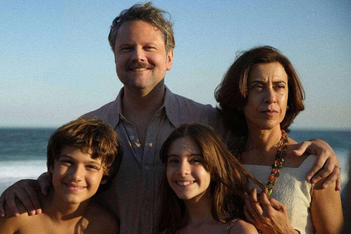 Uma família posando na praia, com o mar ao fundo. O pai, com um sorriso, está no centro, ao lado da mãe, que tem uma expressão séria. À esquerda, um menino sorridente e à direita, uma menina também sorridente. Todos estão vestidos de forma casual, com a luz do sol iluminando a cena.