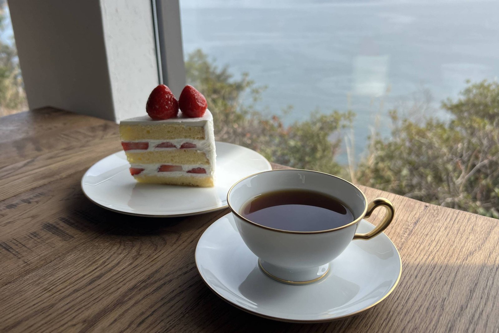 A imagem mostra um pedaço de bolo decorado com morangos em um prato branco, ao lado de uma xícara de café em uma mesa de madeira. Ao fundo, há uma vista de um lago ou mar com ilhas ao longe, sob um céu claro.