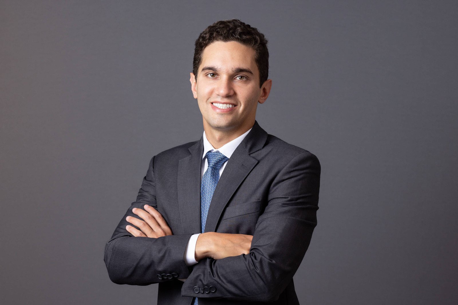 Um homem está posando para a foto, com um sorriso no rosto. Ele está vestindo um terno escuro e uma camisa branca, com uma gravata azul. O fundo da imagem é cinza, e o homem está com os braços cruzados na frente do corpo.