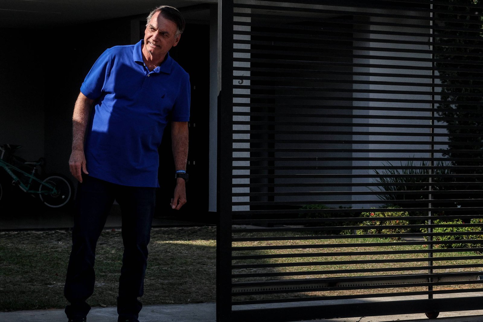 Um homem está em pé em frente a um portão preto, que está parcialmente aberto. Ele usa uma camisa azul e calças escuras, e parece estar olhando para fora. Ao fundo, há uma casa com uma parede clara e um gramado. Um objeto verde, possivelmente uma bicicleta, está visível ao lado do homem.