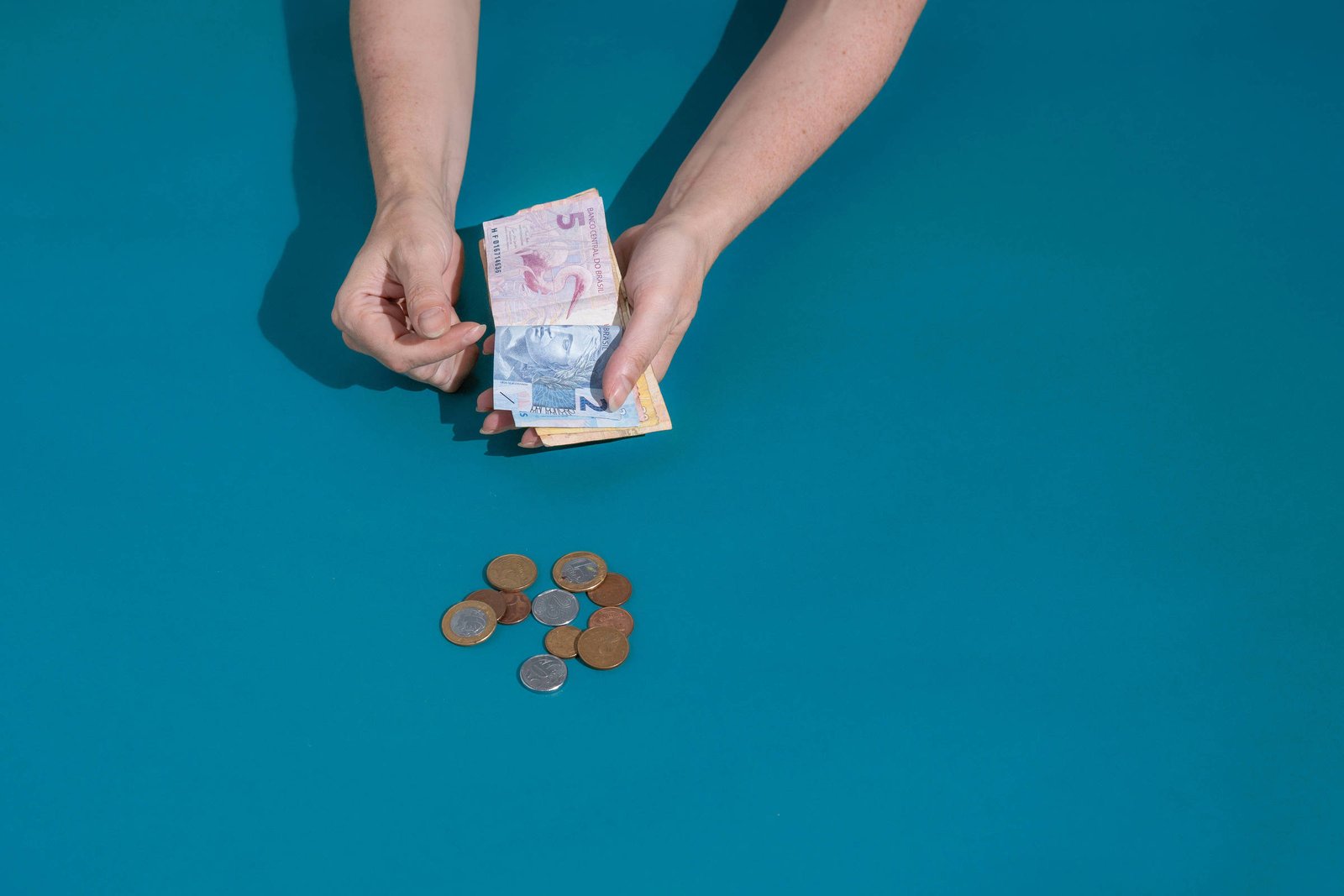 Uma mão segurando notas de dinheiro, enquanto outra mão está próxima a uma pilha de moedas sobre uma superfície azul. As notas incluem uma de 5 e uma de 2, e as moedas estão espalhadas em diferentes tamanhos e cores.