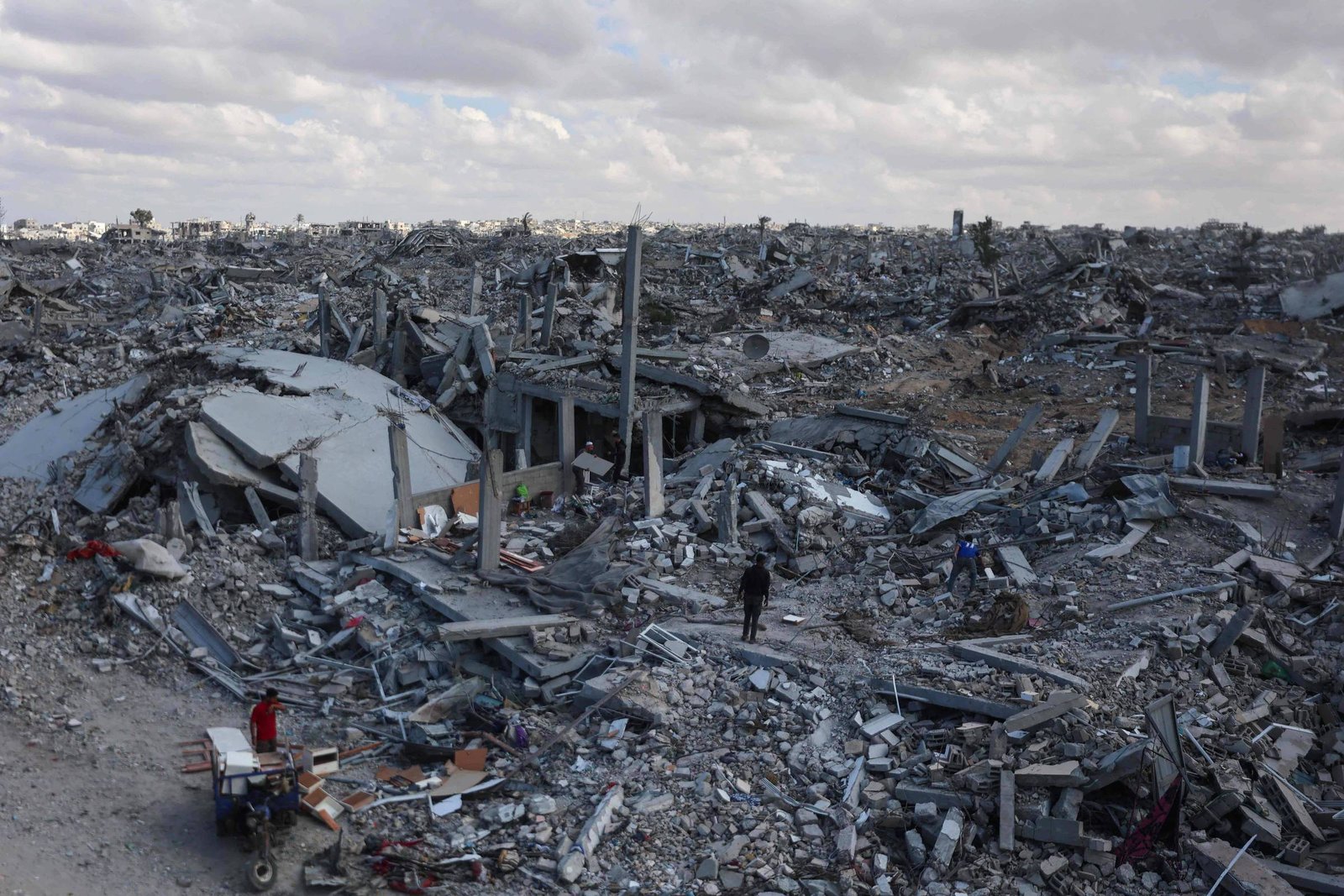 Grande extensão de destroços de edifícios destruídos sob céu nublado. Três pessoas caminham entre os escombros, destacando a escala da destruição.