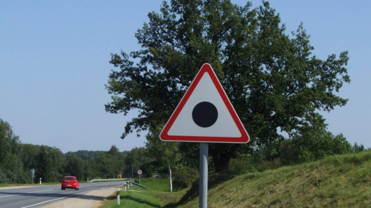 Placa de trânsito com triangulo com bordas vermelhas e ponto preto no meio. Por trás, aparece vegetação e rodovia