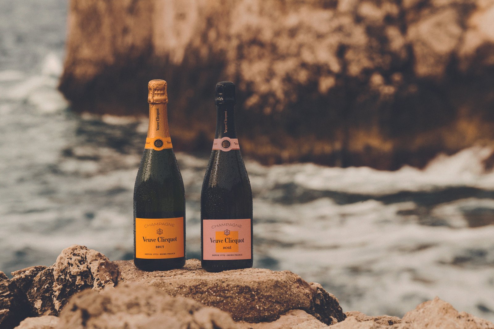 Duas garrafas de champanhe Veuve Clicquot, uma com rótulo amarelo e outra com rótulo rosa, estão posicionadas sobre pedras irregulares próximas ao mar. Ao fundo, ondas quebram contra uma formação rochosa grande e desfocada.