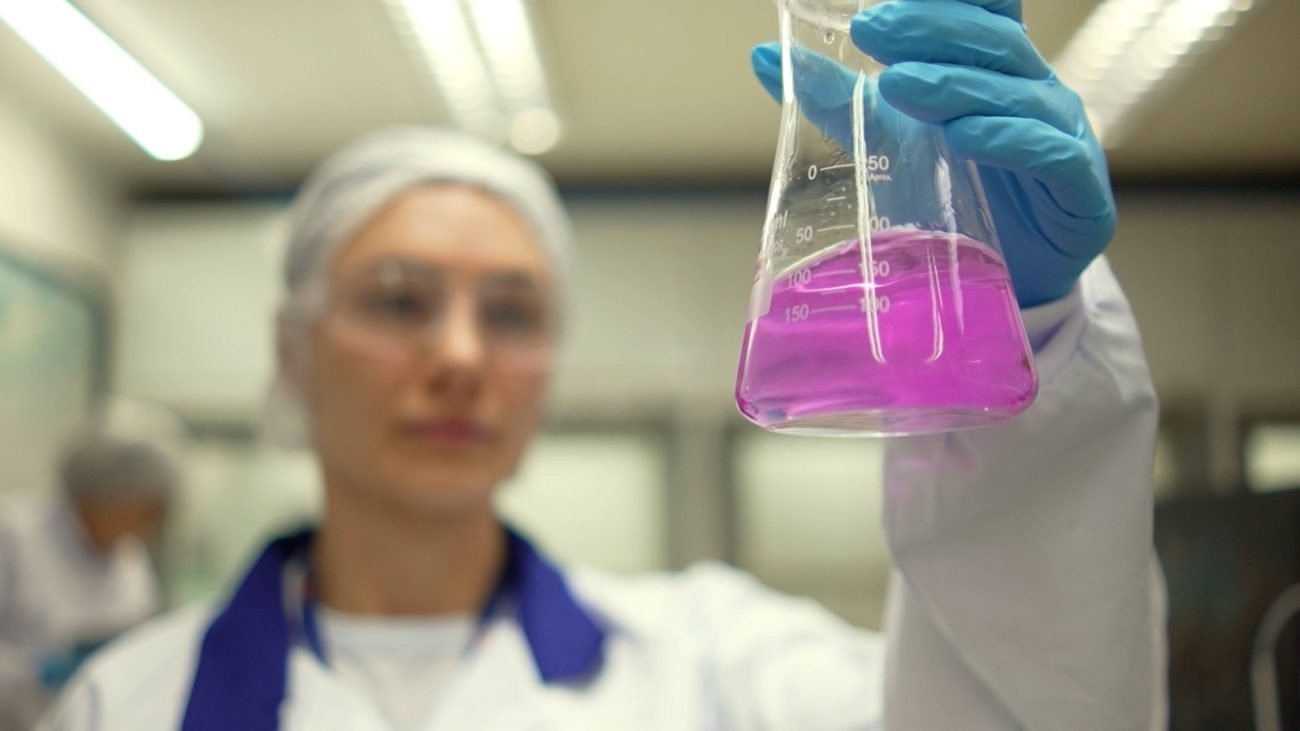 mulher segurando bequer com líquido roxo e roupa branca e luvas azuis, representando pesquisadora impactada por investimento em ciência