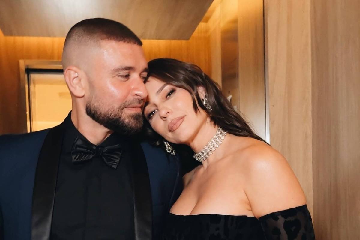 Homem de terno azul escuro com camisa preta e gravata borboleta abraça mulher com vestido preto de ombros à mostra e colar brilhante. Ambos estão próximos, em ambiente com paredes de madeira clara.