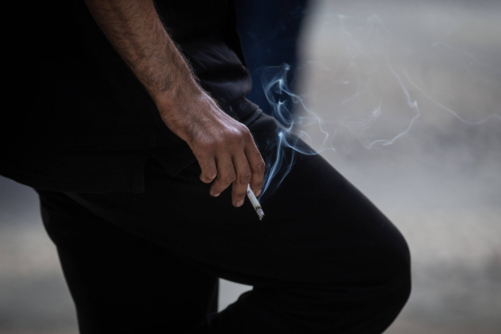 Mão direita de pessoa vestindo roupa preta segura cigarro aceso com fumaça visível. Fundo desfocado em tons neutros.