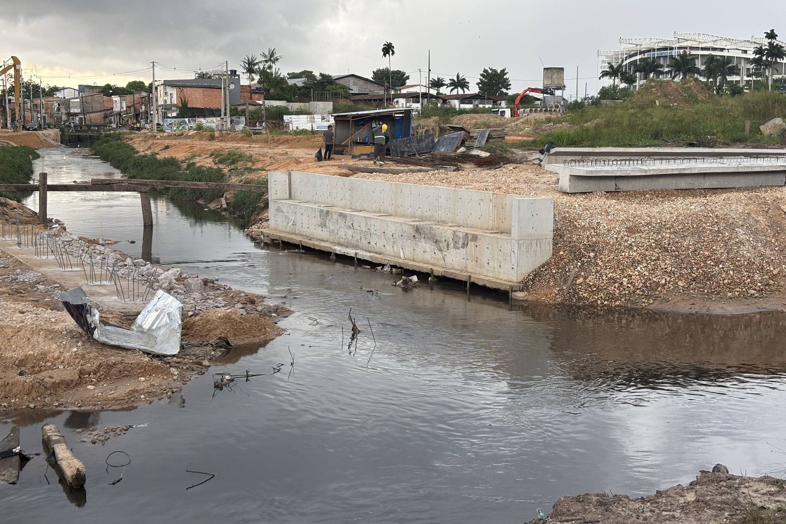 A imagem mostra uma área de construção em um ambiente urbano, com um canal de água escura à esquerda. À direita, há uma estrutura de concreto em construção, com materiais de construção e terra ao redor. No fundo, é possível ver algumas edificações e árvores, além de um céu nublado.