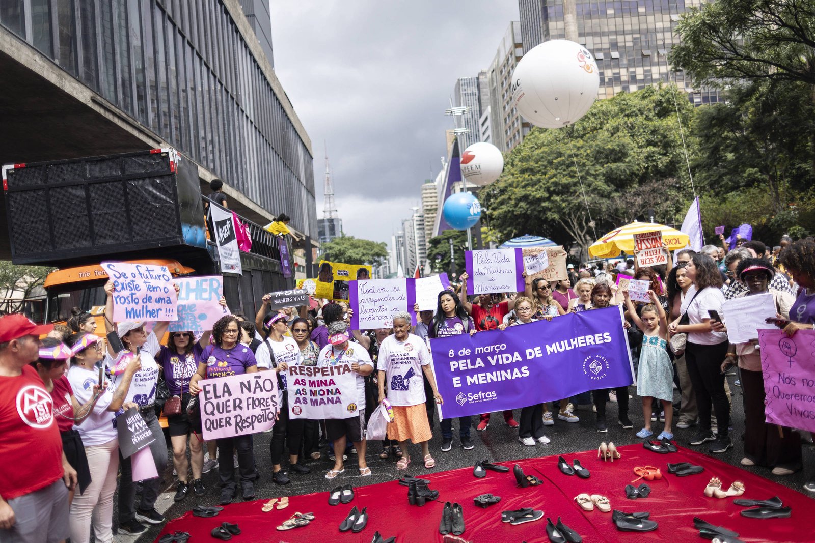 Grupo de pessoas reunidas em manifestação na rua, segurando cartazes e faixa roxa com texto