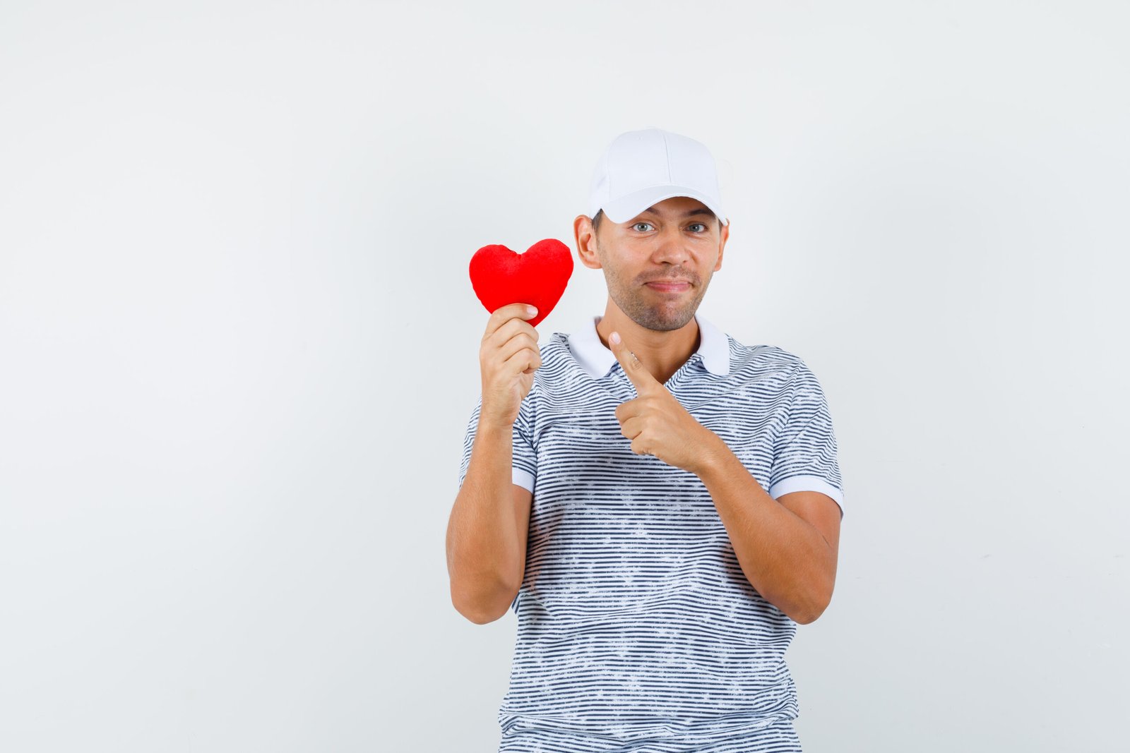 Young male pointing at red heart in t-shirt, cap and looking merry. front view.