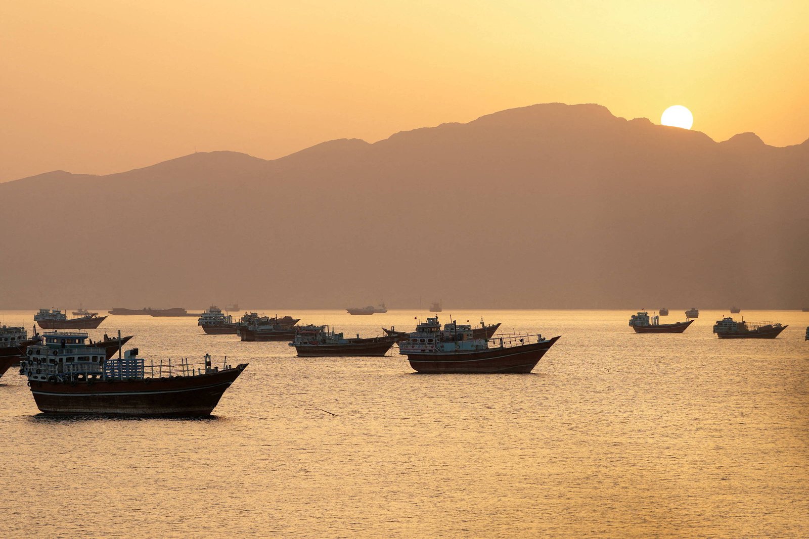 Várias embarcações ancoradas em águas calmas durante o pôr do sol. O sol está parcialmente visível atrás de uma cadeia de montanhas ao fundo, iluminando o céu e a água com tons dourados.