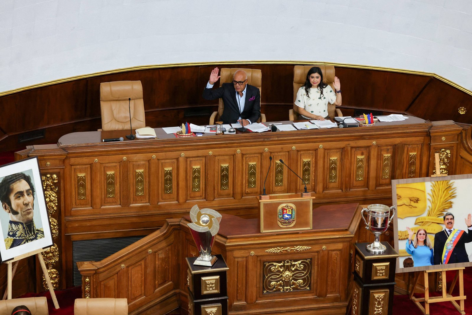 Dois parlamentares sentados em cadeiras altas atrás de bancada de madeira ornamentada em plenário. Ambos com a mão direita levantada, indicando voto. À esquerda, retrato emoldurado de figura histórica; à direita, foto emoldurada de casal com faixa presidencial. Troféus expostos em pedestal de madeira abaixo da bancada.