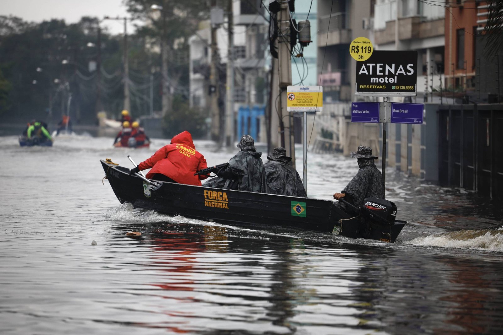 Três pessoas em barco de resgate navegam por rua inundada. Edifícios e placas comerciais visíveis ao fundo em dia nublado.