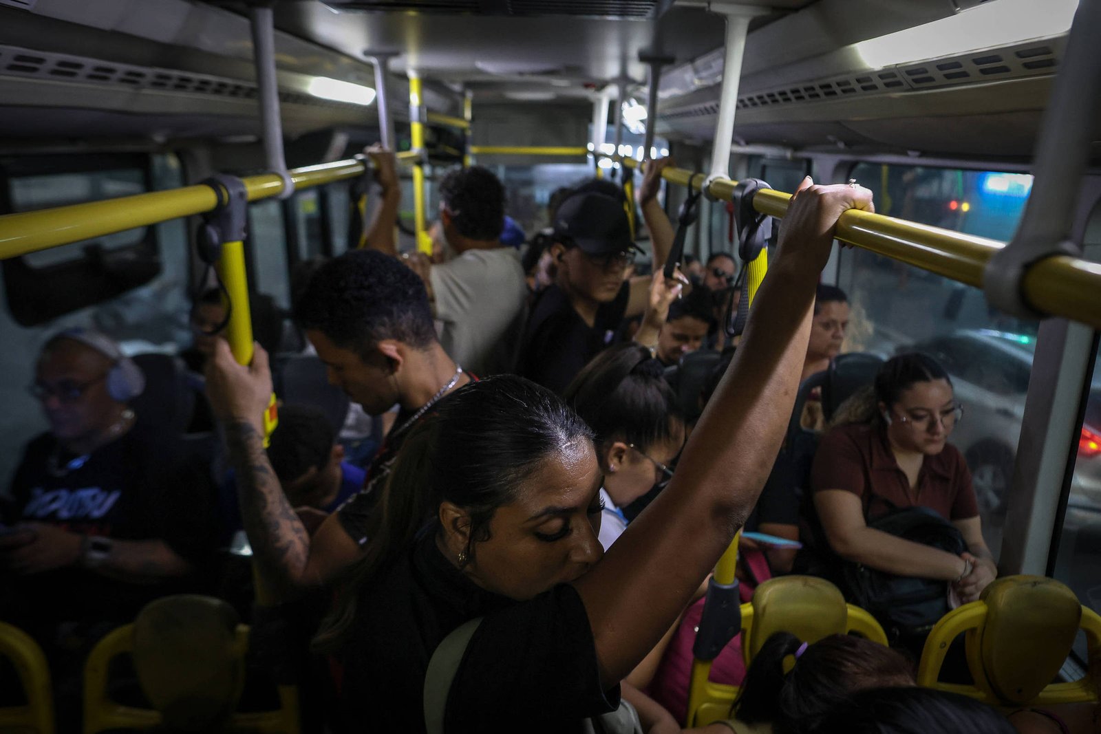 Diversos passageiros estão em pé e sentados dentro de um ônibus urbano lotado à noite. Alguns seguram os suportes amarelos no teto enquanto outros estão sentados, alguns usando fones de ouvido. Luzes internas iluminam o espaço apertado.