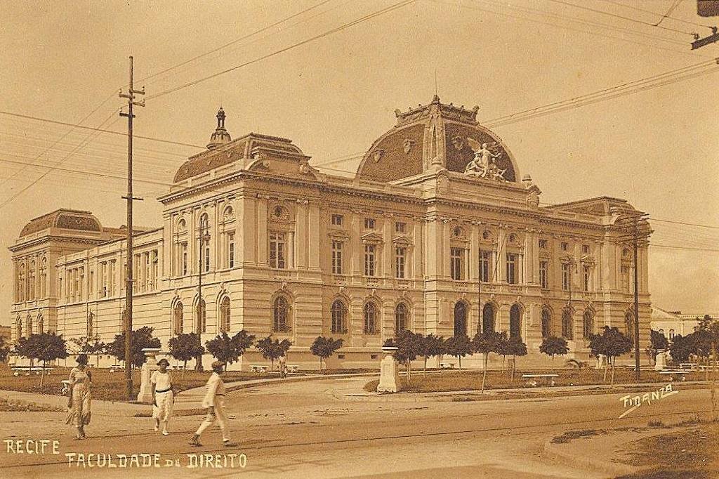 Edifício clássico da Faculdade de Direito em Recife com arquitetura neoclássica, janelas altas e detalhes ornamentais. Três pessoas caminham na calçada em frente, árvores pequenas alinham a rua e postes de eletricidade com fios cruzam o céu.