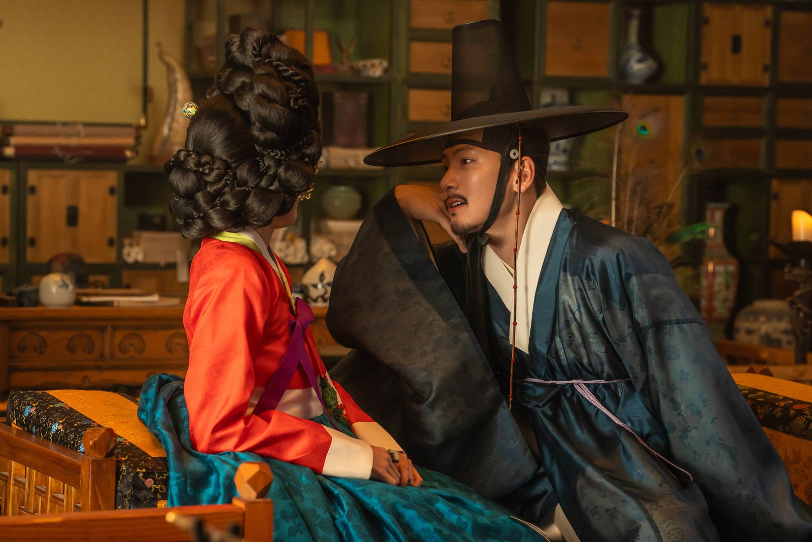 Homem e mulher vestidos com hanbok tradicional coreano sentados em móveis de madeira entalhada. O homem usa chapéu gat e veste roupa azul escura, enquanto a mulher usa vestido vermelho e azul com penteado elaborado. Ambiente interno com móveis antigos e objetos decorativos ao fundo.