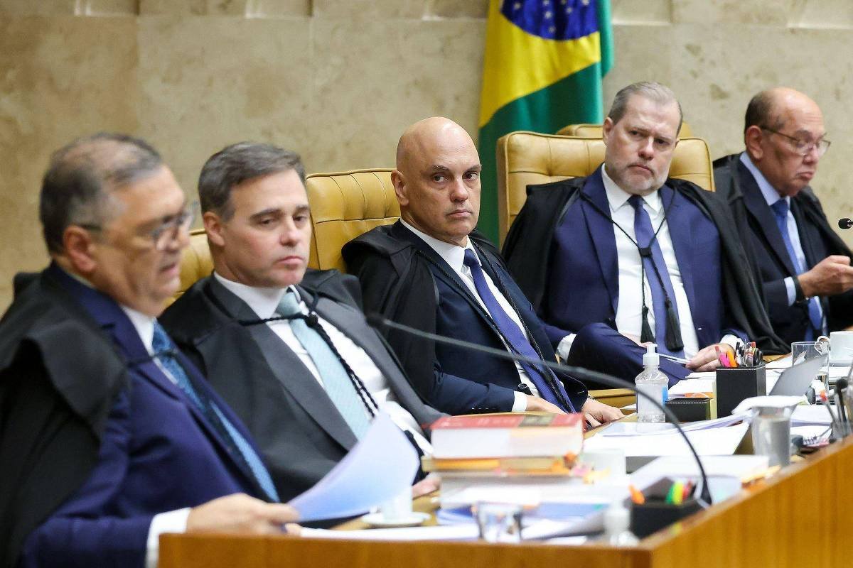 Cinco ministros do Supremo Tribunal Federal sentados em bancada durante sessão. Ao fundo, bandeira do Brasil. Documentos e microfones estão sobre a mesa.