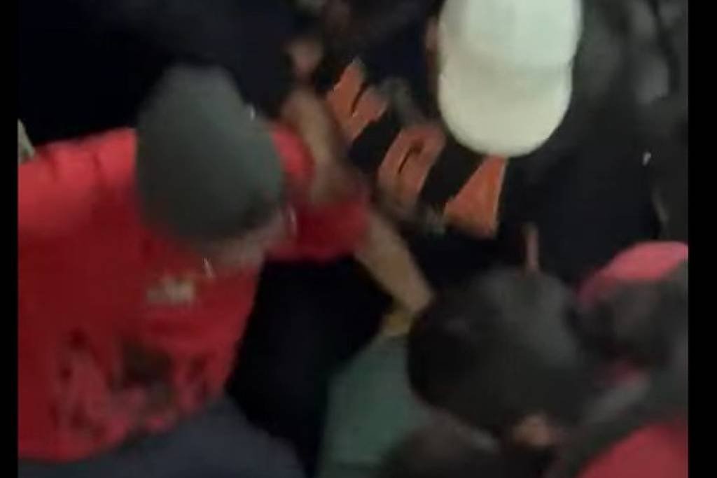 Cinco pessoas cercam e seguram um indivíduo vestido de preto no chão. Uma pessoa usa boné branco e outra veste camiseta vermelha. Ambiente interno com pouca iluminação.