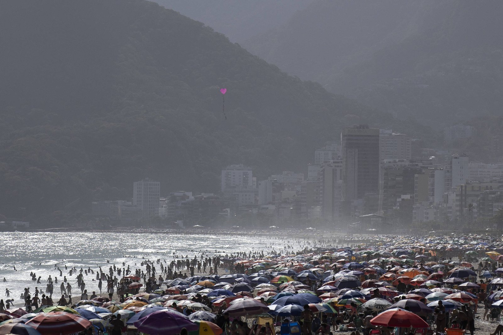 Multidão de pessoas na areia e no mar de uma praia urbana com muitos guarda-sóis coloridos. Ao fundo, prédios altos e morros cobertos por vegetação sob céu nublado.