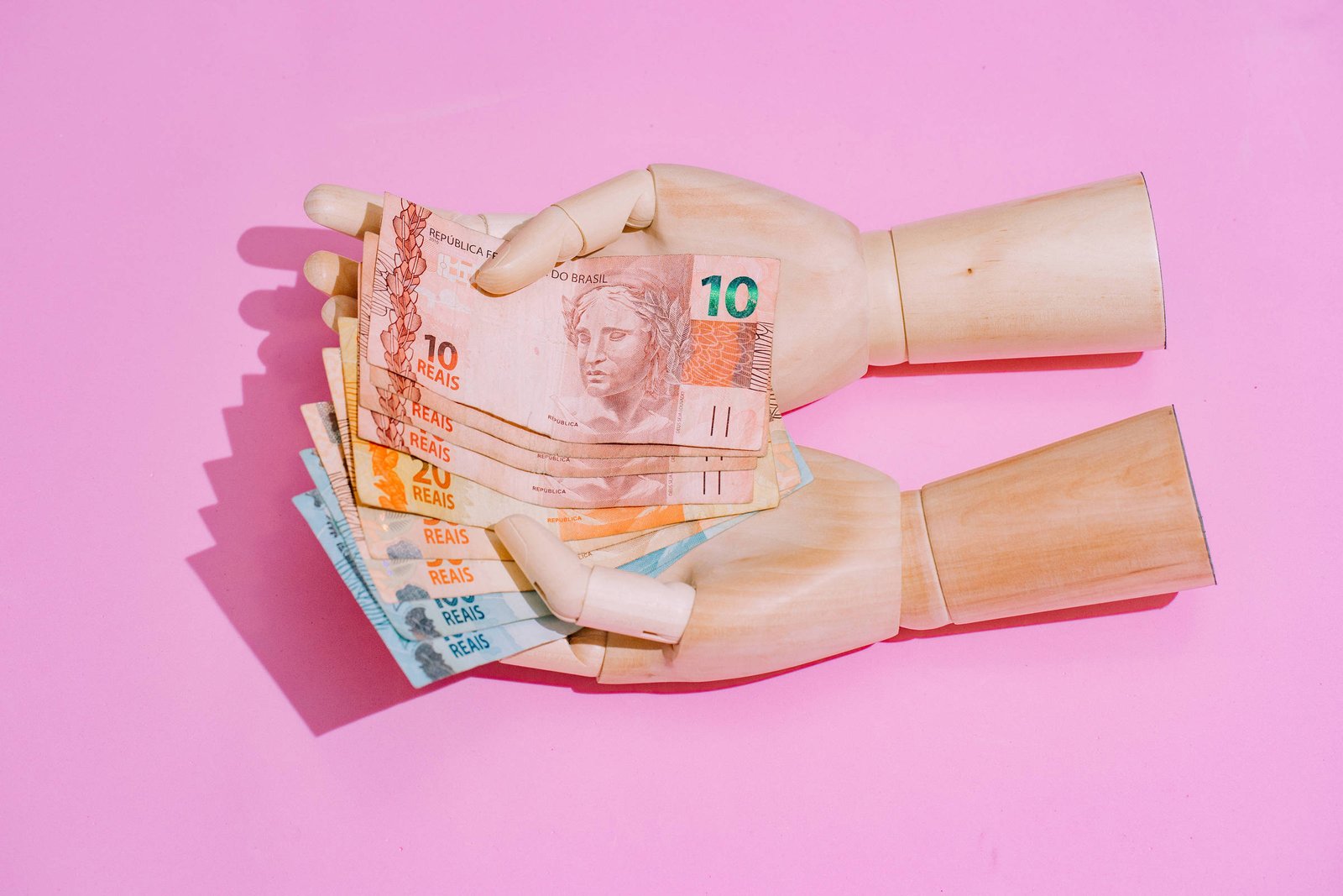 Duas mãos de madeira articuladas seguram várias cédulas de reais brasileiros em diferentes valores, sobre fundo rosa.