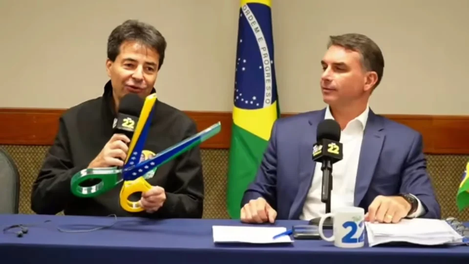 Sachsida segura tesoura com as cores da bandeira do Brasil que simboliza promessa de corte de gastos em live ao lado de Flávio Bolsonaro (PL)